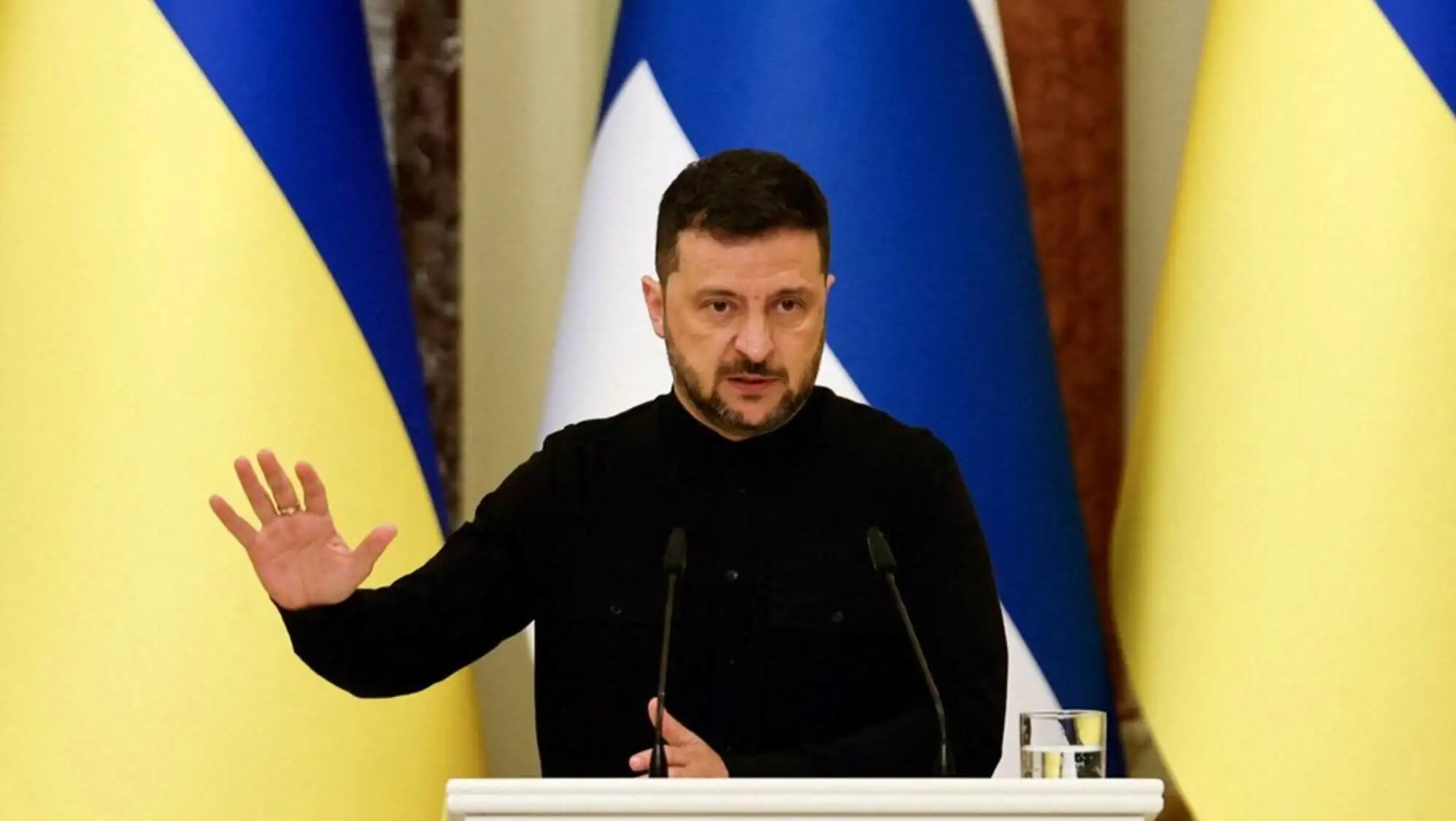 Bir adam mikrofonla bir podiumda duruyor, arkasında bir bayrak var.
