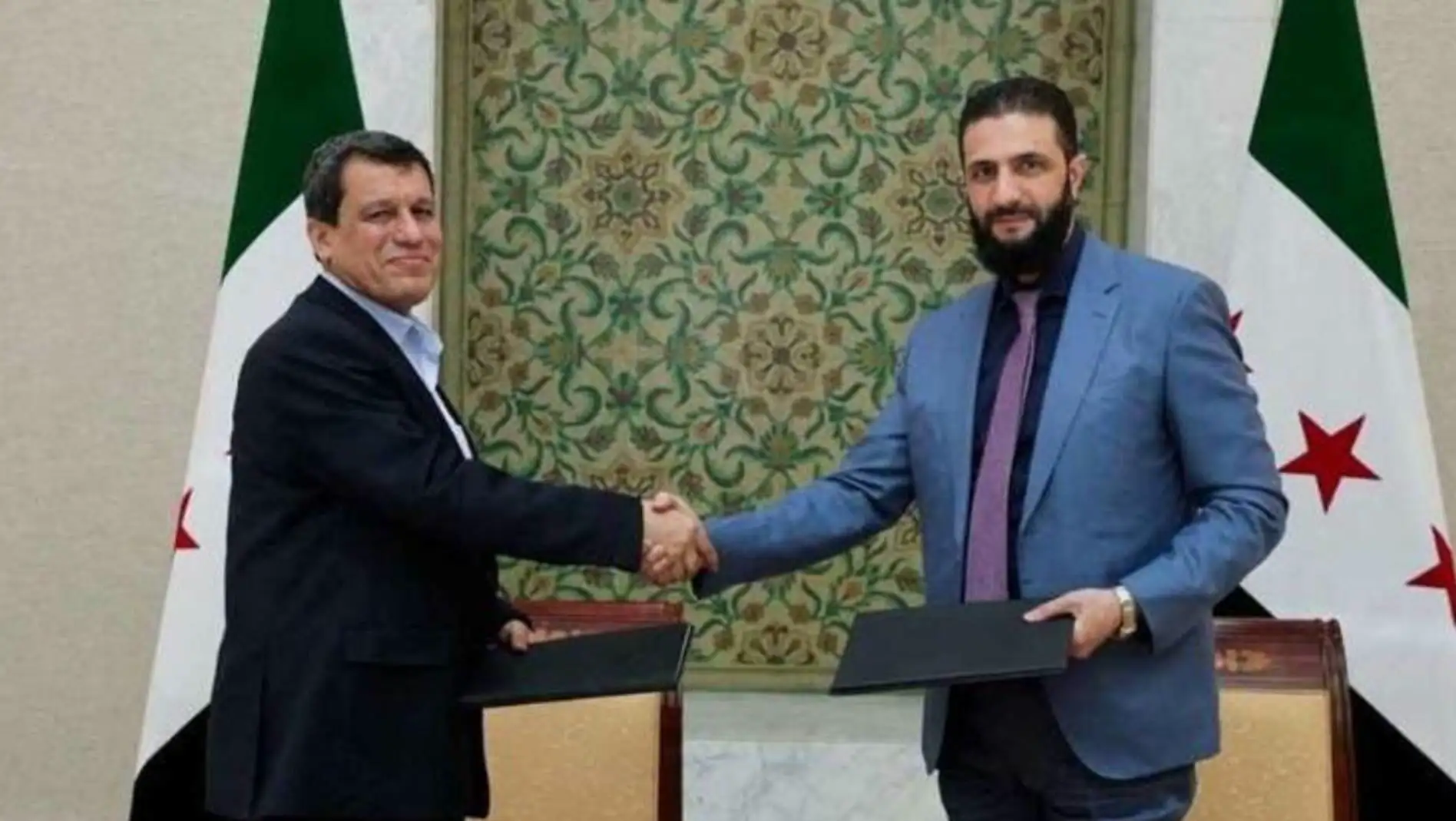 Two men shaking hands in front of a green wall.
