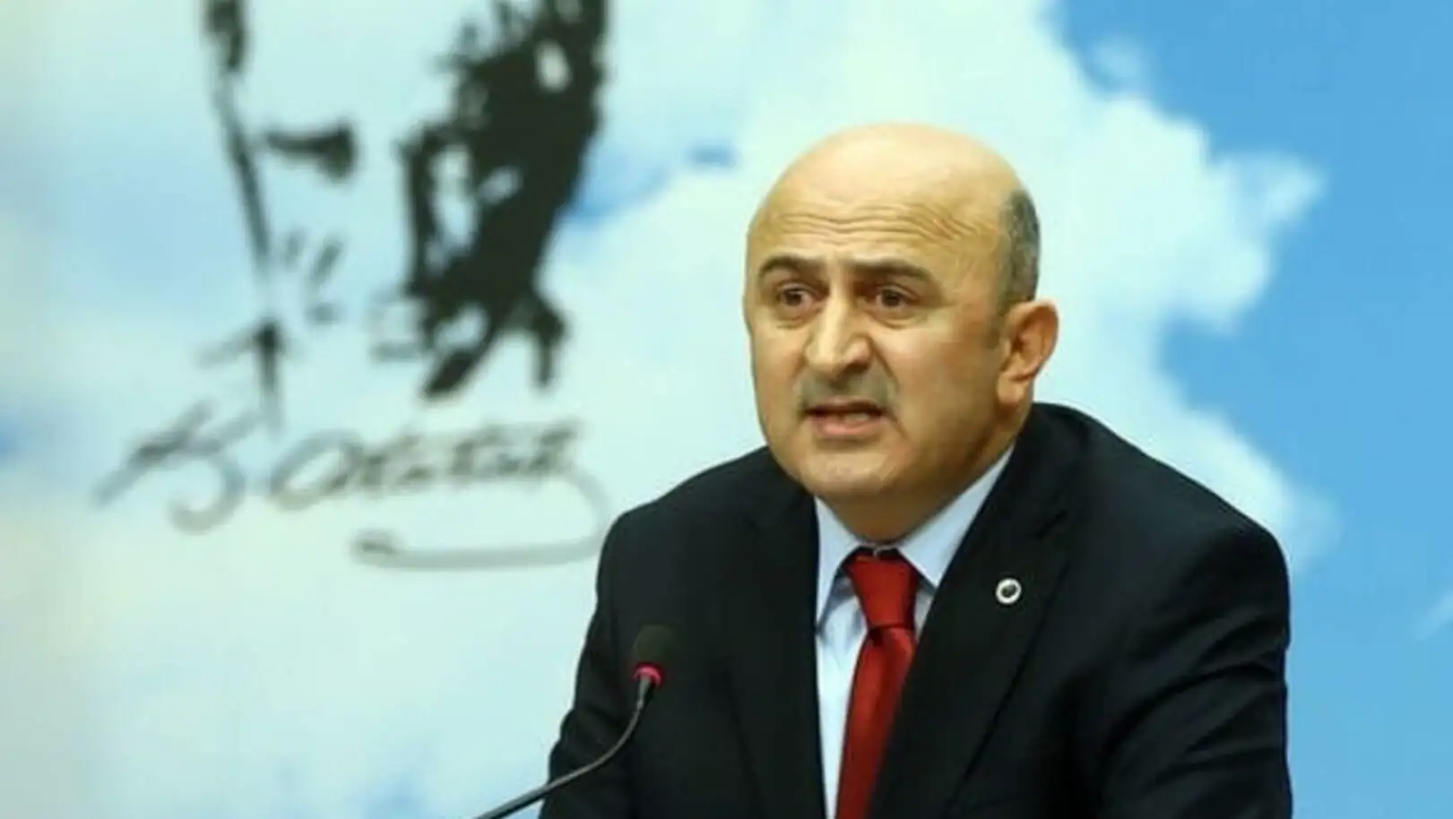 Bir adam takım elbise giymiş ve mikrofona konuşuyor.